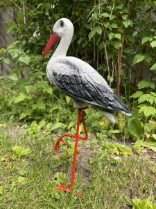 Storchfigur stehend mit roten Beinen in natuerlicher Gartenumgebung.